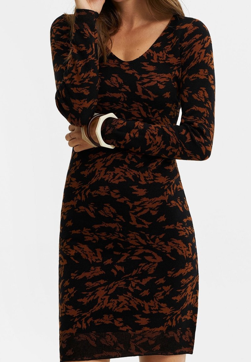 Femme portant une robe en tricot à manches longues à motifs abstraits noirs et bruns avec un décolleté en V, accessoirisée de bracelets épais blancs et bruns.