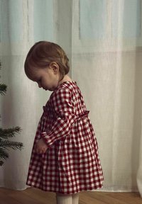 Vestido a cuadros rojos y blancos con mangas largas, cintura fruncida y detalle de botones en la parte posterior. Un niño está de pie junto a un árbol, con una textura suave en la tela.