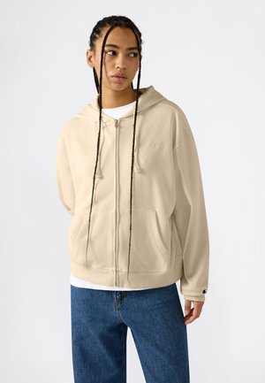 Jeune personne aux longs cheveux tressés portant un sweat à capuche beige zippé et un jean bleu, regardant de côté sur un fond uni.