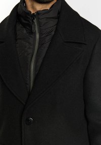 Abrigo negro de doble botonadura con amplias solapas sobre una chaqueta acolchada con cremallera. Presenta una tela texturizada, costuras prominentes y un acabado mate.
