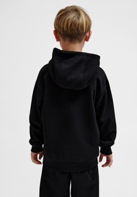 Svart polotröja med en rymlig huva, långa ärmar, ribbade mudd och fåll. Slät textur, avslappnad passform, bärs med matchande svarta byxor.