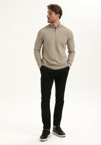 Pulóver beige de canalé con cuello de cremallera y mangas largas, combinado con pantalones negros ajustados y zapatillas negras con suelas blancas.