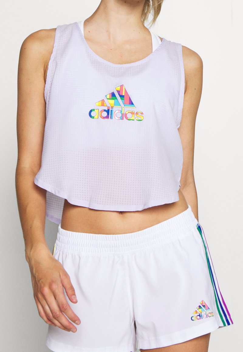 Weißes, kurz geschnittenes sportliches Tanktop mit strukturiertem Muster und mehrfarbigem Adidas-Logo. Kombiniert mit weißen Shorts, die Regenbogen-Seitenstreifen haben.
