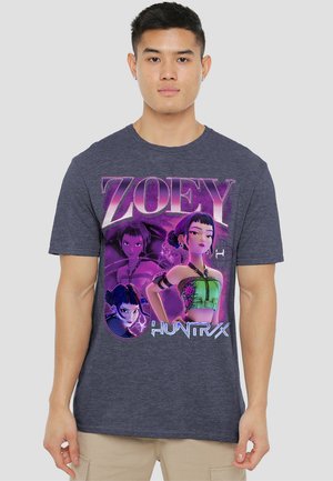 Young man wearing a dark gray shirt with colorful animated female character "Zoey" from Huntrix in purple and green graphic design.
