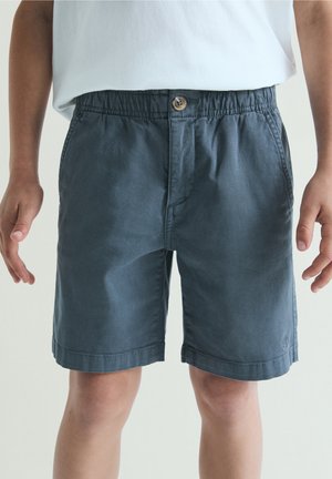 Niño con pantalones cortos elásticos de cintura oscura gris y una camisa blanca, de pie con los brazos relajados a los lados.