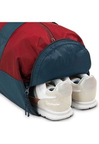 Bolsa de deporte con paneles rojos y azules, que presenta un compartimento con cremallera que contiene zapatillas blancas con acentos en beige y plata.