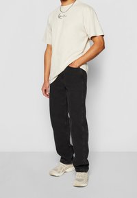 T-shirt beige in cotone con logo in script nero, abbinato a jeans neri e sneakers beige, che mostrano una vestibilità rilassata e un design casual.
