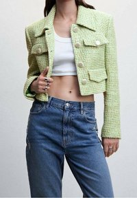 Donna che indossa una giacca corta in tweed verde sopra un crop top bianco e jeans blu a vita alta, mostrando la pancia e la mano con anelli d'argento.