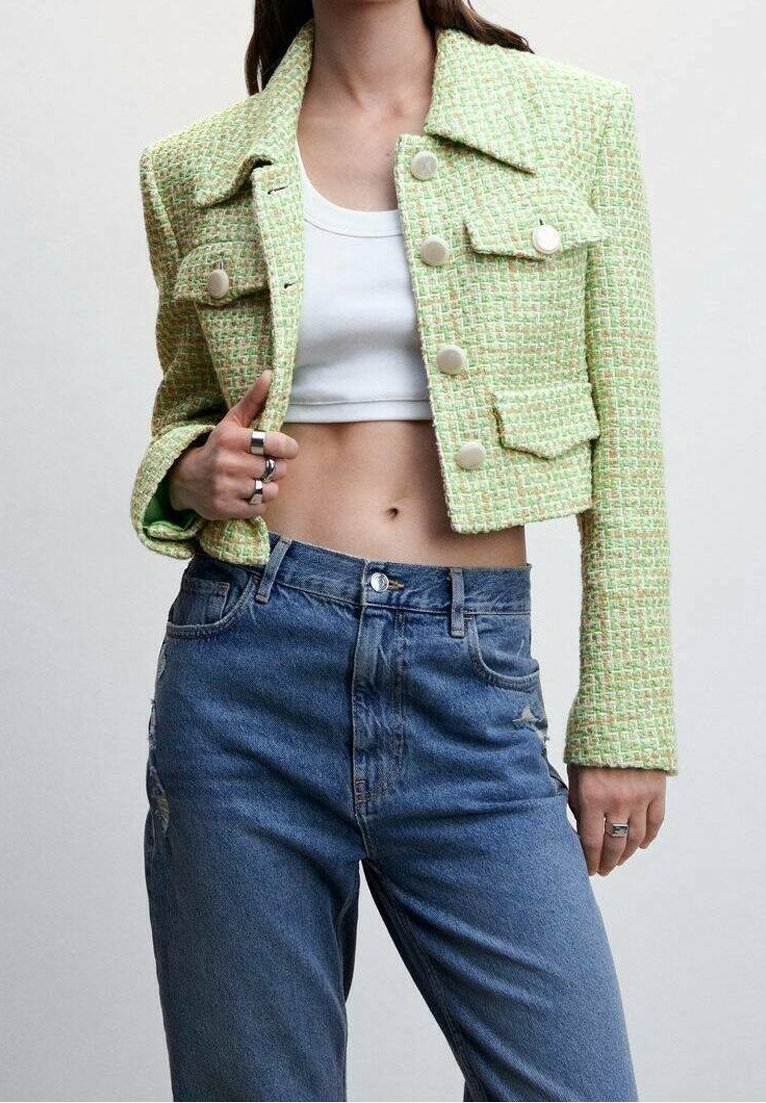 Donna che indossa una giacca corta in tweed verde sopra un crop top bianco e jeans blu a vita alta, mostrando la pancia e la mano con anelli d'argento.