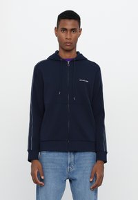 Felpa con zip di colore blu navy realizzata in tessuto morbido, con cappuccio regolabile, strisce laterali e un piccolo logo bianco sul petto.
