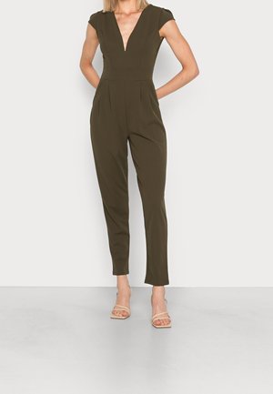 Kvinde iført en olivengrøn ærmeløs jumpsuit med dyb V-udskæring og beige åbne hælede sandaler, stående med hænderne bag ryggen.