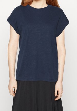 Nainen, joka käyttää löysää tummansinistä lyhythihaista t-paitaa ja mustaa hametta, kuvattuna kaulasta lantioihin.