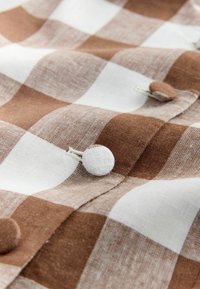 Hnědá a bílá kostkovaná látka s potaženými knoflíky přišitými na oděvu, zobrazující detaily stehování a textury.