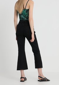 Jean noir évasé avec taille haute et ourlet brut, associé à un haut vert à motifs et des sandales noires ornées de clous et d'une boucle.