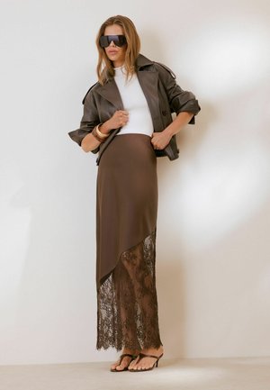 Kvinde iført store solbriller, en cropped brun læderjakke, hvid top, brun asymmetrisk nederdel med blonderkant og sorte hælede sandaler.
