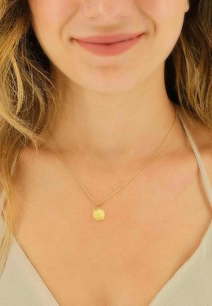 Collier en chaîne dorée avec un design torsadé et un pendentif rond arborant un motif en relief, porté sur un haut de couleur claire.