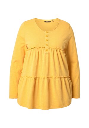 Keltainen pitkähihais Toppi, joka on valmistettu kevyestä kankaasta. Siinä on pyöreä kaula-aukko, nappilista ja kerroksellinen, rypytetty helma.