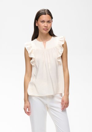 Femme debout, portant un chemisier crème sans manches à épaules à volants et un pantalon blanc, sur un fond uni clair.