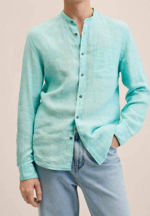 Mann trägt ein helltürkisfarbenes Leinenlangarmhemd mit Mandarin-Kragen, schwarzen Knöpfen, Brusttasche und hellblauen Jeans, Hand in der Tasche.