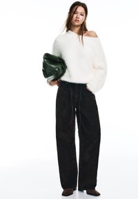 Młoda kobieta w białym, puszystym swetrze z odkrytymi ramionami, luźnych czarnych spodniach, brązowych butach, trzymająca dużą ciemnozieloną kopertówkę.