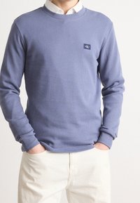 Man som bär en ljuslila ribbad tröja över en vit skjorta, med händerna i fickorna, iförd krämfärgade byxor framför en enkel ljusbakgrund.