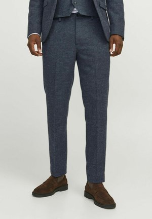 Mann trägt maßgeschneiderte marineblaue Anzugshosen und passenden Blazer mit braunen Wildleder-Loafers, steht vor einem schlichten hellen Hintergrund.