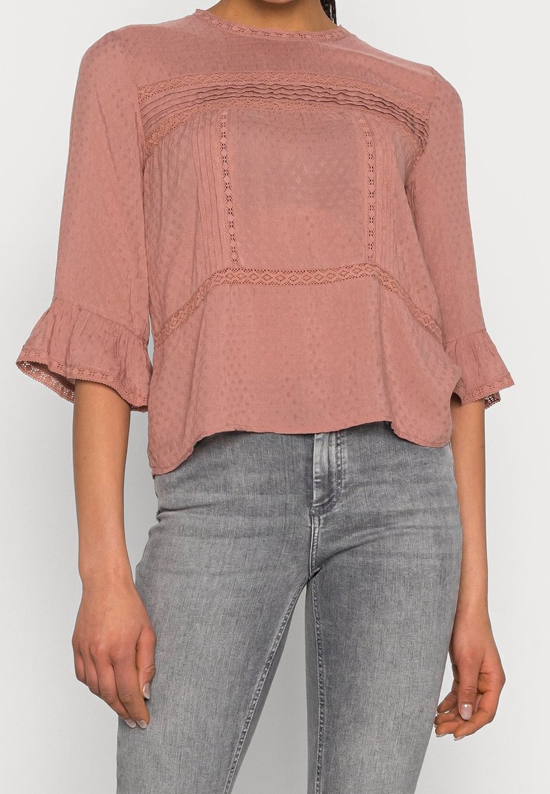 Femme portant un chemisier texturé rose poudré avec des poignets à volants et un jean gris clair ajusté, présenté des épaules aux hanches.
