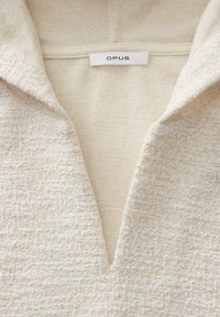 Textured cream-colored garment with a V-neck collar and subtle sheen. Features a label with the brand name "OPUS" visible at the neckline.