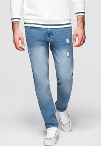 Jean en denim bleu clair avec des empiècements usés, coupe slim, design à cinq poches et lavage délavé. Associé à des baskets blanches.