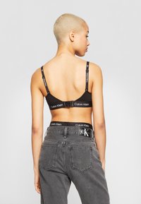 Svarte Calvin Klein bralette med justerbare stropper og logo-bånd. Modellen har på seg grå høytlivsjeans med logo-bånd i livet og baklommer.