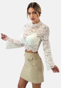 Witte kanten crop top met lange wijde mouwen, gecombineerd met een beige mini-rok met een decoratieve knoop en ritsdetail.