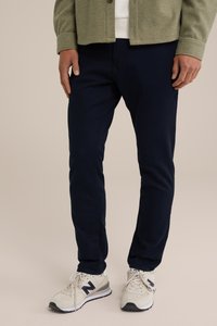 Tamne plave tapered hlače s glatkom teksturom, uparene s bež New Balance tenisicama s tamnim logom i bijelim akcentima.