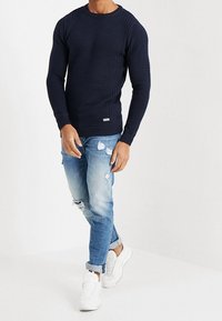 Man in een marineblauwe geribbelde lange mouw trui, gescheurde lichtblauwe opgerolde jeans en witte sneakers, staand tegen een plain witte achtergrond.
