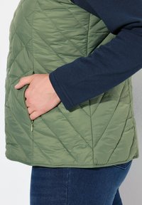 Zelený prošívaný vest s diagonálním švem, s boční kapsou na zip, nošený přes námořnickou košili s dlouhým rukávem a modré džíny.