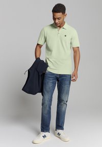 Lichtgroene polo met korte mouwen, voorzien van een kraag en knoopdetails. Te combineren met donkere jeans en witte sneakers. Marineblauwe jas in de hand gehouden.