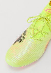 Botín de fútbol de color verde lima con parte superior sintética texturizada, acentos reflectantes, detalles de patrón en olas negros y agujetas en contraste de neón.