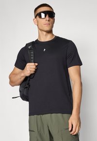 Sort kortærmet t-shirt med rund halsudskæring og et lille hvidt logo på brystet. Modellen bærer en sort rygsæk og har mørke solbriller på.