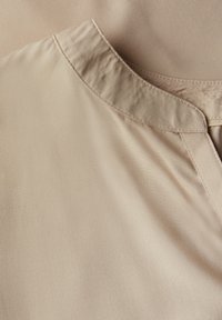 Beige Kleidungsstück mit glatter Textur, das über einen geschwungenen Ausschnitt und gefaltete Kanten verfügt. Der Stoff wirkt leicht und elegant.