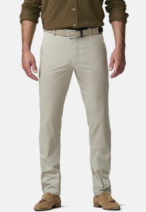 Homme portant un pantalon beige slim, une ceinture tressée claire, une chemise marron boutonnée avec les manches retroussées, et des mocassins à glands en daim couleur sable, debout.
