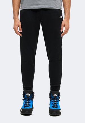 Parte inferior del cuerpo de una persona que lleva pantalones negros North Face y botas de senderismo negras y azul brillante con cordones y logotipo sobre un fondo claro.