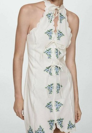Kobieta ubrana w białą sukienkę na szyję bez rękawów z falowanymi krawędziami, niebieską kwiatową haftem i trzema wiązaniami z przodu.