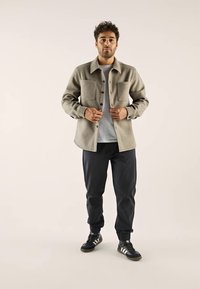 Homme debout portant une veste beige à boutons sur une chemise gris clair, un pantalon de jogging noir et des baskets noires avec des rayures blanches.