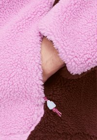 Luddig rosa fleecetyg med en plyschliknande textur, med en brun insida och ett dekorativt dragsnöre med en vit pärla som accent.