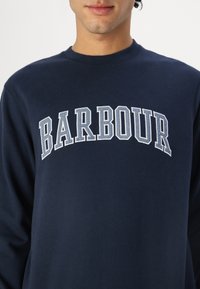 Σκουρό μπλε φανελάκι βαμβακερό με τη λέξη "BARBOUR" σε αψιδωτά, ανοιχτόχρωμα γράμματα κατά μήκος του στήθους. Λείος υφασμάτινος κόμπος, λαιμόκοψη τύπου crew.
