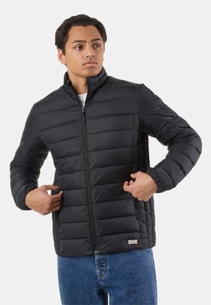 Giovane uomo che indossa una giacca trapuntata nera e jeans blu, con le mani che tengono i lati della giacca vicino alle tasche, guarda verso sinistra.