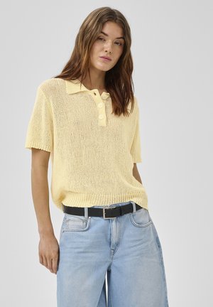 Młoda kobieta ubrana w jasnożółtą, dzianą, krótkim rękawem koszulkę polo, włożoną do jasnoniebieskich dżinsów z czarnym paskiem.