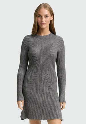 Femme portant une robe ajustée en maille côtelée grise à manches longues avec un col rond, debout devant un fond clair uni.