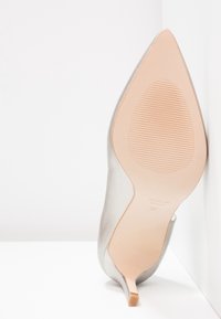 Chaussure à talons aiguilles argentée avec un bout pointu, texture en cuir lisse et talon fin en stiletto. La semelle est de couleur beige clair avec des détails en relief.