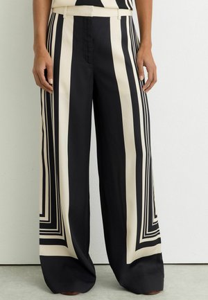 Sorte bukser med vide ben og markante, lodrette hvide og sorte geometriske striber, båret af en person, der står op mod en ensfarvet væg.