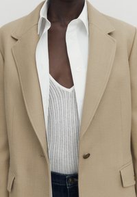 Blazer beige su misura con revers a notched sopra una camicia bianca e un gilet grigio a coste. Dettagli notevoli includono i bottoni e una vestibilità strutturata.